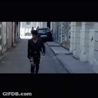A Man Is Walking Down A Street With A Skateboard In His Hand GIF