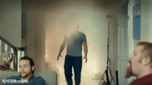 A Man Is Walking Through A Doorway With Smoke Coming Out Of It While A Man Sits On A Couch . GIF