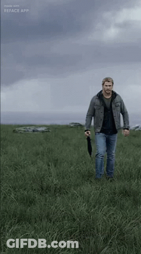 A Man Is Walking Through A Grassy Field With An Umbrella GIF