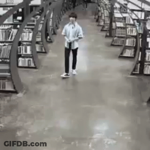 A Man Is Walking Through A Library Filled With Bookshelves . GIF