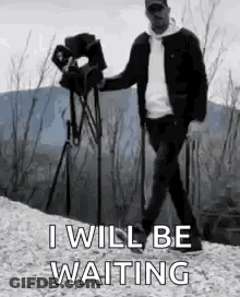 A Man Is Walking With A Camera On A Tripod While Holding A Camera . GIF