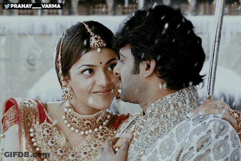 A Man Kisses A Woman On The Cheek In Front Of A Sign That Says Pranay_varma GIF