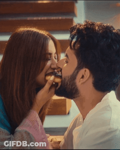A Man Kisses A Woman On The Cheek While She Eats A Doughnut GIF