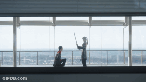 A Man Kneeling Down With A Woman Holding A Stick In Front Of A Large Window GIF