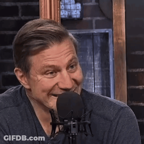 A Man Laughs In Front Of A Microphone With A Brick Wall Behind Him GIF