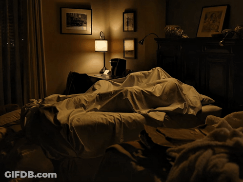 A Man Laying On A Bed In A Dark Room With A Lamp On GIF