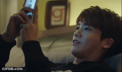 A Man Laying On A Bed Looking At A Cell Phone With The Letter G On The Wall Behind Him GIF