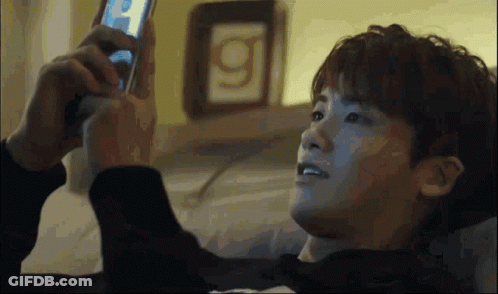 A Man Laying On A Bed Looking At A Cell Phone With The Letter G On The Wall Behind Him GIF