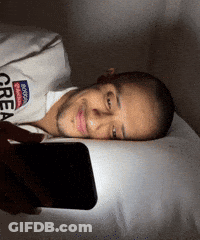 A Man Laying On A Bed Looking At His Phone With A Shirt That Says Brooklyn Great GIF