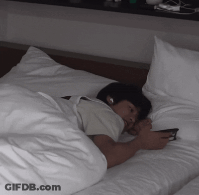 A Man Laying On A Bed With White Sheets Looking At His Phone GIF