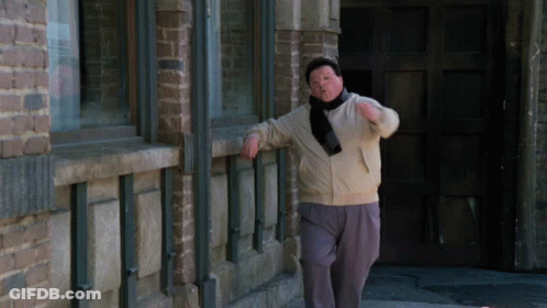 A Man Leans Against A Brick Wall While Wearing A Scarf Around His Neck GIF