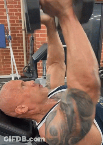 A Man Lifting A Dumbbell In A Gym With A Brick Wall In The Background GIF