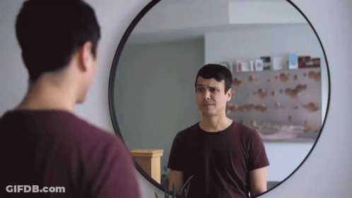 A Man Looks At Himself In A Round Mirror With A Reflection Of A Shelf With Books On It GIF