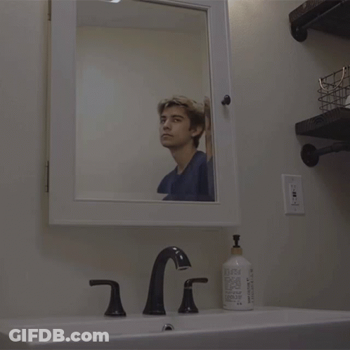 A Man Looks At His Reflection In A Mirror In A Bathroom GIF
