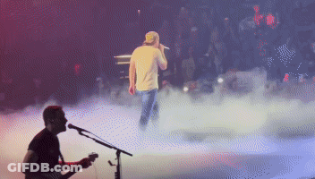 A Man Playing A Guitar And Singing Into A Microphone On A Stage GIF