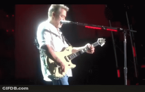 A Man Playing A Guitar And Singing Into A Microphone On A Stage GIF
