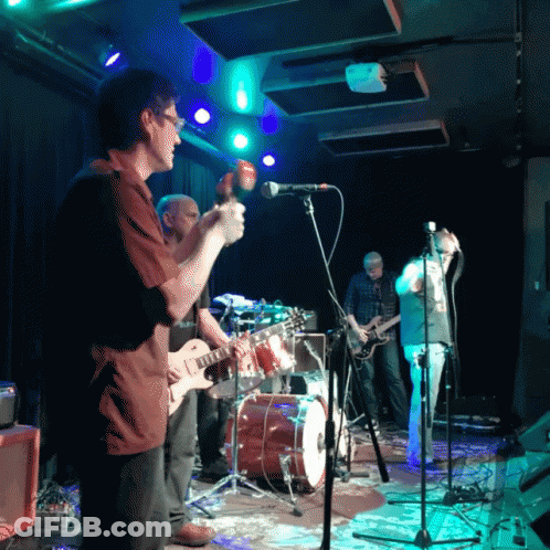 A Man Playing A Guitar Stands In Front Of A Microphone GIF