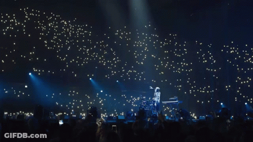 A Man Playing A Keyboard On A Stage With A Crowd Behind Him GIF