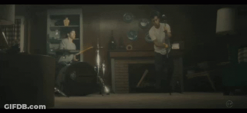 A Man Playing Drums In A Living Room With A Man Playing A Guitar In The Background GIF