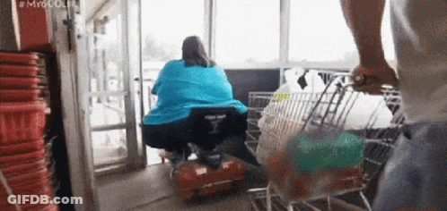 A Man Pushes A Shopping Cart With A Woman In A Wheelchair In The Background GIF