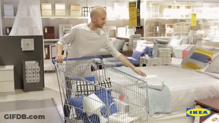 A Man Pushing A Shopping Cart In An Ikea Store With A Bed In The Background GIF