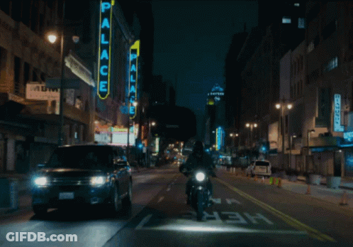 A Man Riding A Motorcycle Down A City Street With A Neon Sign That Says Palace GIF