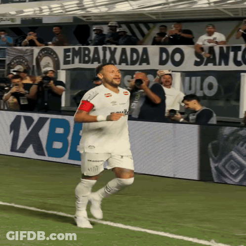 A Man Running On A Soccer Field With A Banner Behind Him That Says " Embaixada Do Santos " GIF