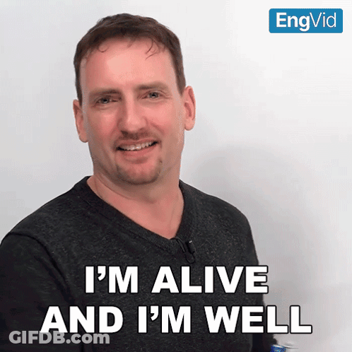 A Man Says " I 'M Alive And I 'M Well " In Front Of A White Wall GIF