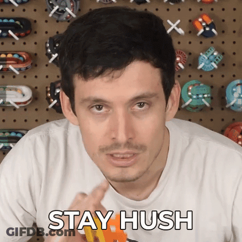 A Man Says " Stay Hush " In Front Of A Wall Of Toys GIF