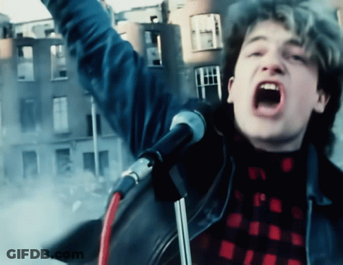 A Man Singing Into A Microphone In Front Of A Building GIF