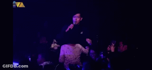 A Man Singing Into A Microphone In Front Of A Crowd In A Dark Room GIF