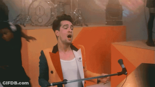 A Man Singing Into A Microphone In Front Of A Drum Set With An Exclamation Point On It GIF