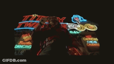 A Man Singing Into A Microphone In Front Of A Lit Up Sign That Says Titty Twist GIF