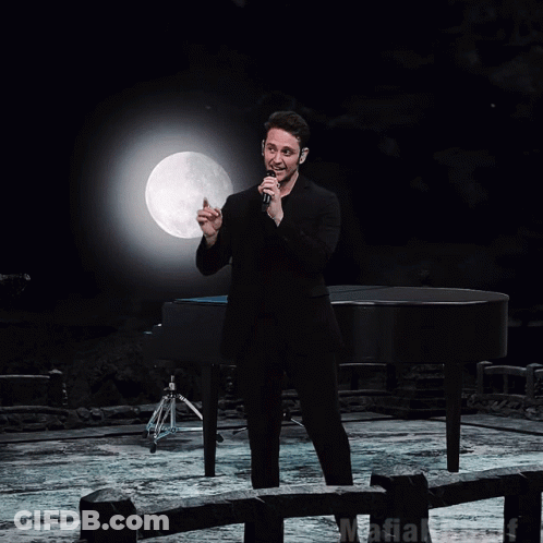 A Man Singing Into A Microphone In Front Of A Piano GIF