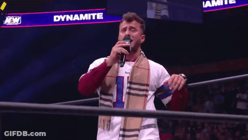 A Man Singing Into A Microphone In Front Of A Sign That Says Dynamite On It GIF