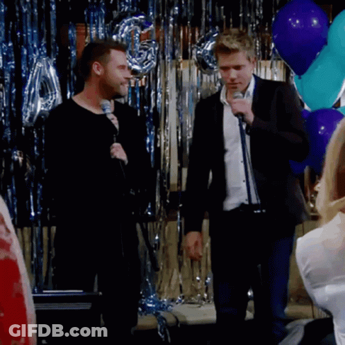 A Man Singing Into A Microphone In Front Of Balloons And Tinsel GIF
