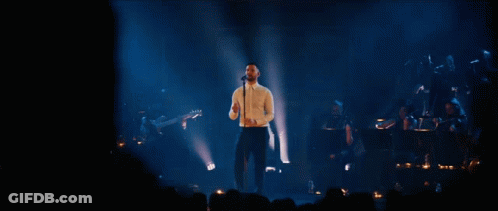 A Man Singing Into A Microphone On A Stage In Front Of A Crowd GIF
