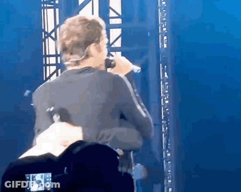 A Man Singing Into A Microphone On A Stage With A Camera Behind Him . GIF