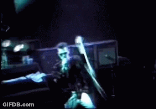 A Man Singing Into A Microphone On A Stage With A Light Behind Him GIF