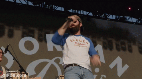 A Man Singing Into A Microphone Wearing A Shirt That Says Serger Customs GIF