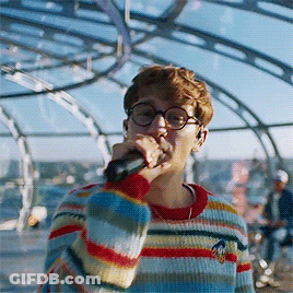 A Man Singing Into A Microphone Wearing A Striped Sweater GIF