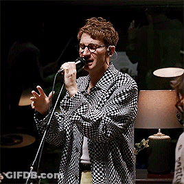 A Man Singing Into A Microphone Wearing Glasses And A Plaid Shirt GIF