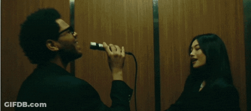 A Man Singing Into A Microphone While A Woman Looks On GIF