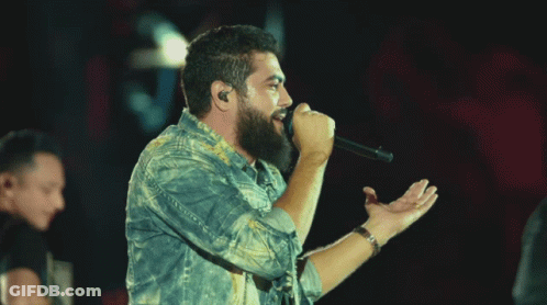 A Man Singing Into A Microphone While Another Man Plays A Guitar In Front Of A Crowd GIF