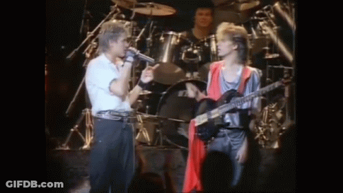 A Man Singing Into A Microphone While Another Man Plays A Guitar On Stage . GIF