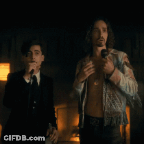 A Man Singing Into A Microphone While Another Man Stands Behind Him GIF