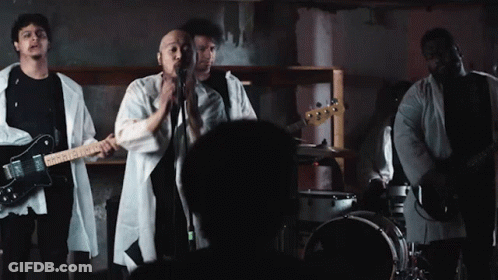 A Man Singing Into A Microphone While Playing A Guitar And A Drum Set GIF