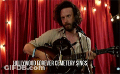A Man Singing Into A Microphone While Playing A Guitar With The Words Hollywood Forever Cemetery Sings Written Below Him GIF