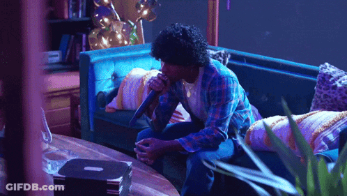 A Man Singing Into A Microphone While Sitting On A Blue Couch GIF
