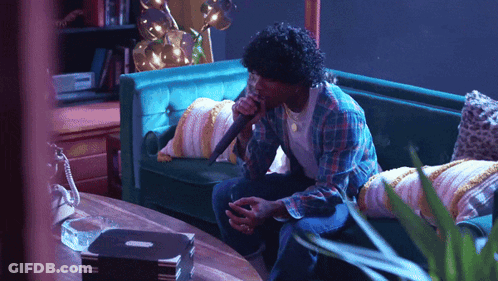A Man Singing Into A Microphone While Sitting On A Green Couch GIF